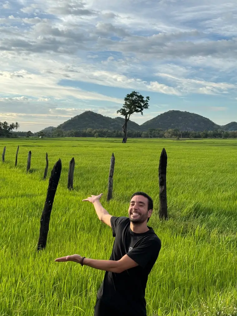 Mahmoud smiling in a green rice field with hills behind, hands held out as if presenting something